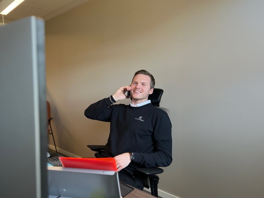 Medarbetare i Hellbergs-tröja sitter vid sitt skrivbord och pratar i mobiltelefon